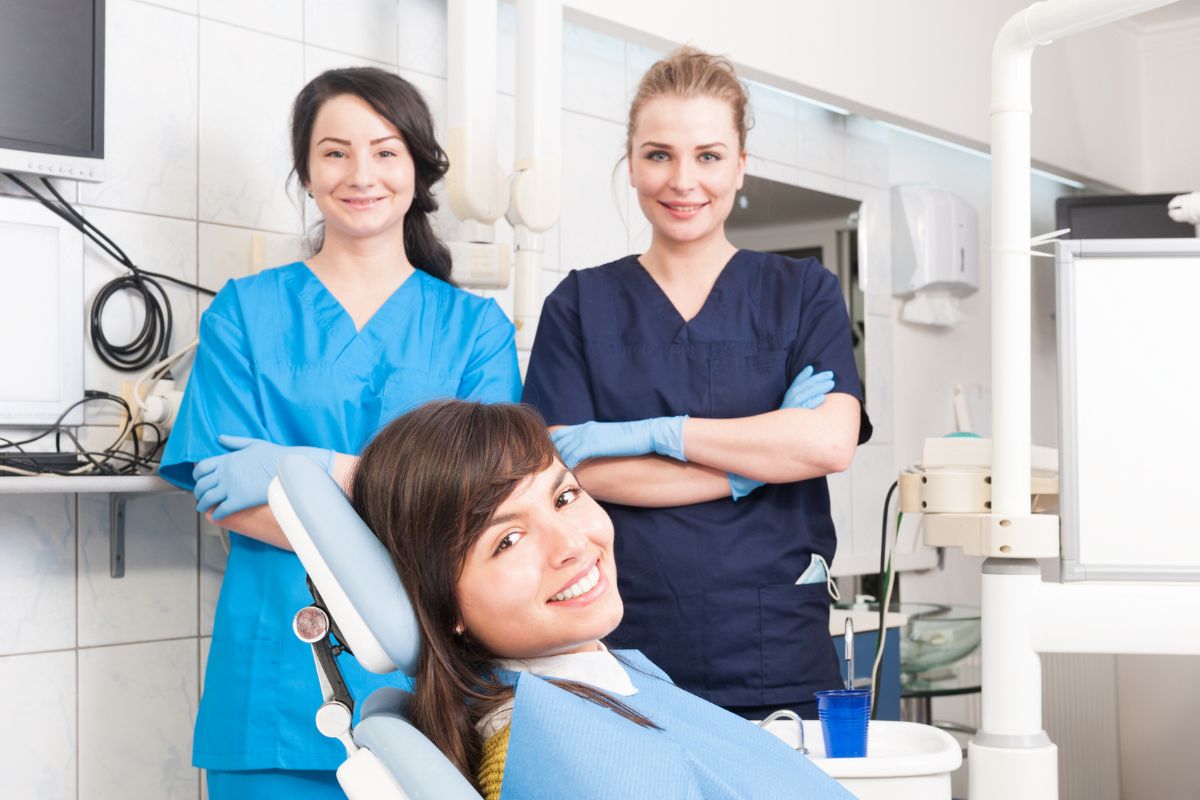 Whats the Difference Between a Dentist and a Hygienist Two female dental professionals stand behind a patient in a modern dental operatory.